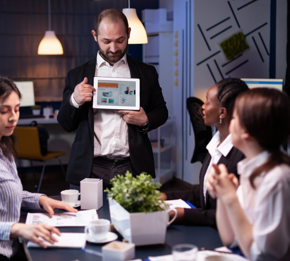 Focused businessman showing corporate graphs presentation using tablet working at company ideas. Diverse multi-ethnic businesspeople brainstorming strategy late at night in office meeting room